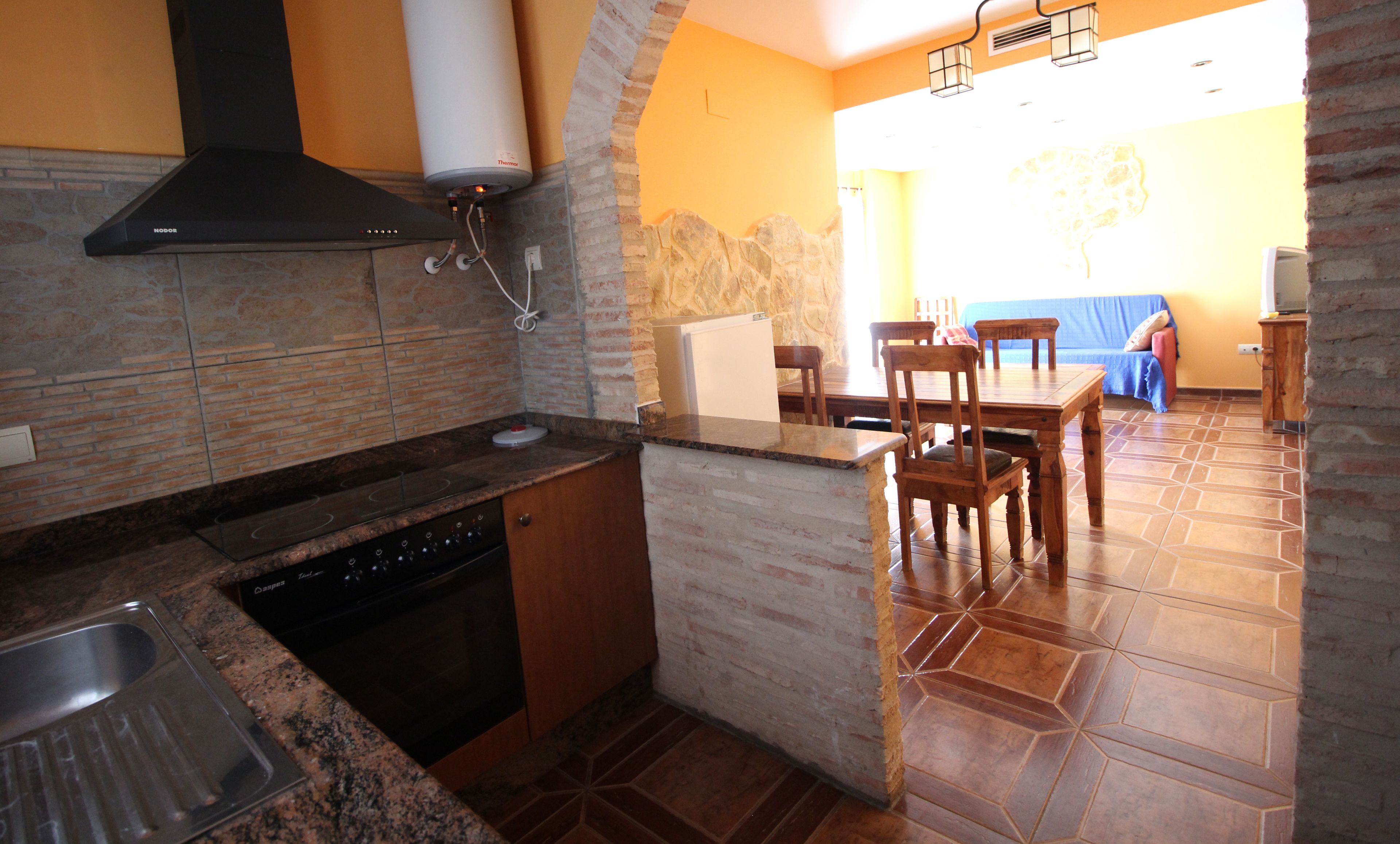 Una cocina con estufa, lavavajillas y calentador de agua. Hay una zona de comedor con mesa y sillas.