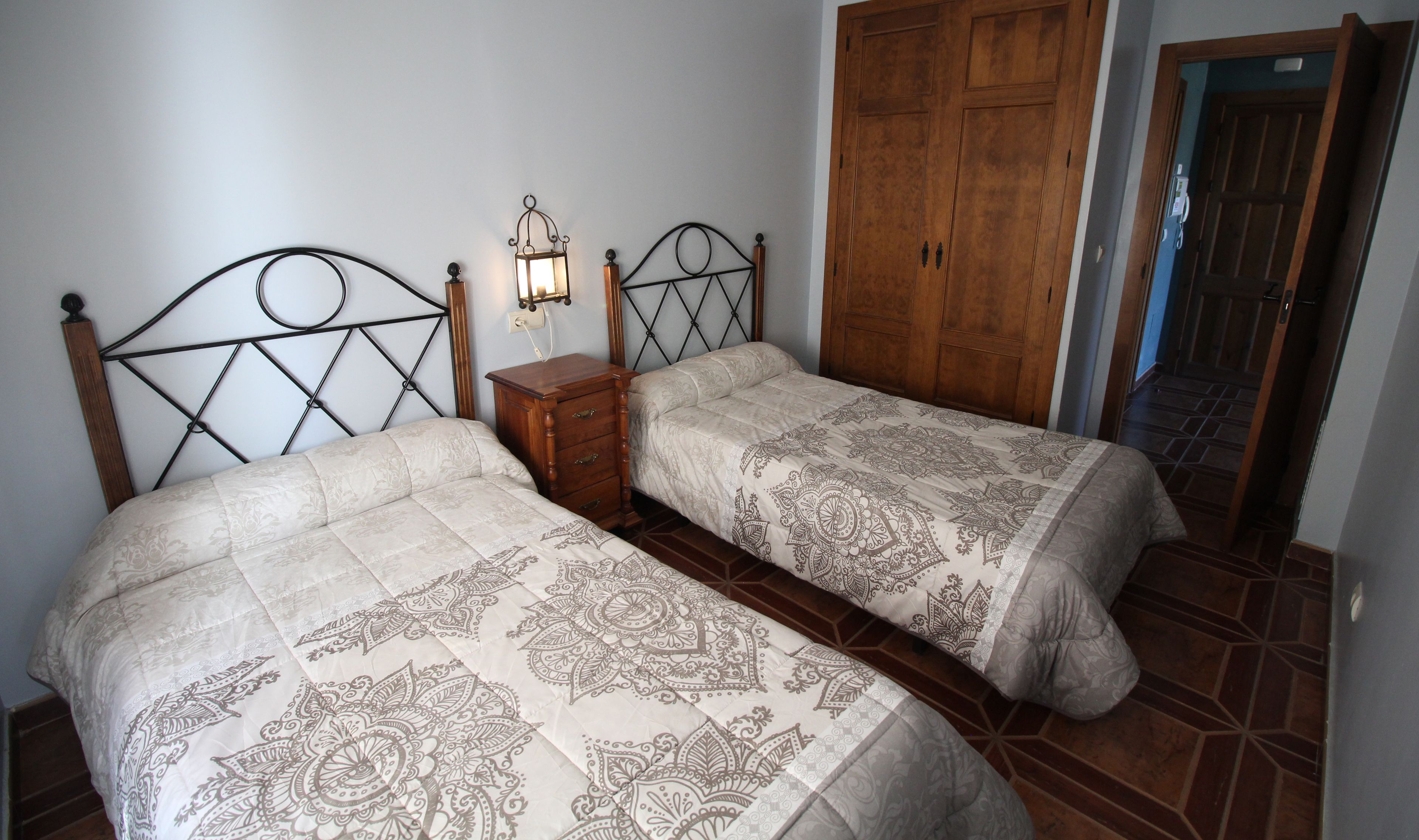 A bedroom with two beds, a wooden wardrobe, and a nightstand with a lamp.