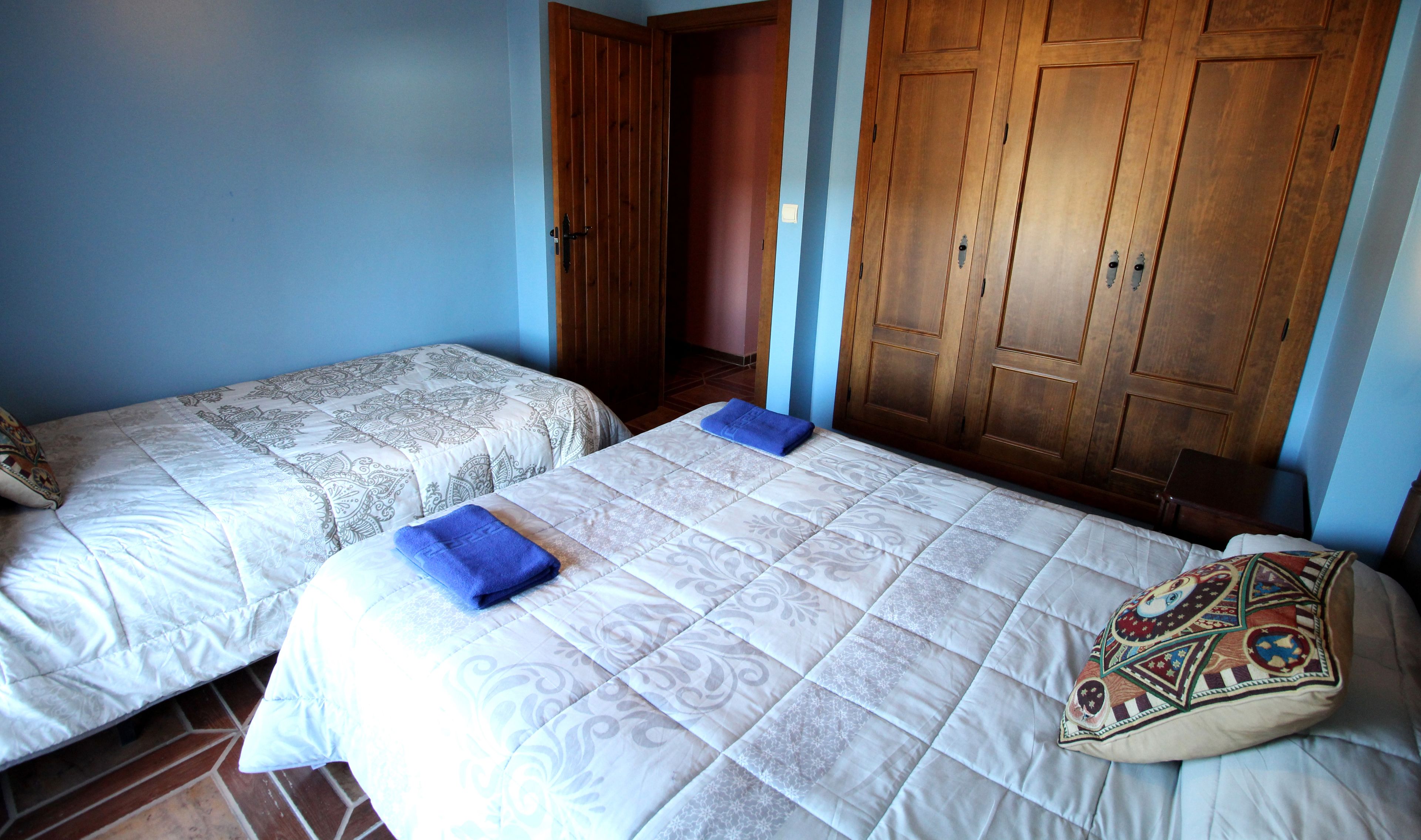 Two single beds with patterned bedding and a blue towel, wooden wardrobe, and a door leading to another room.