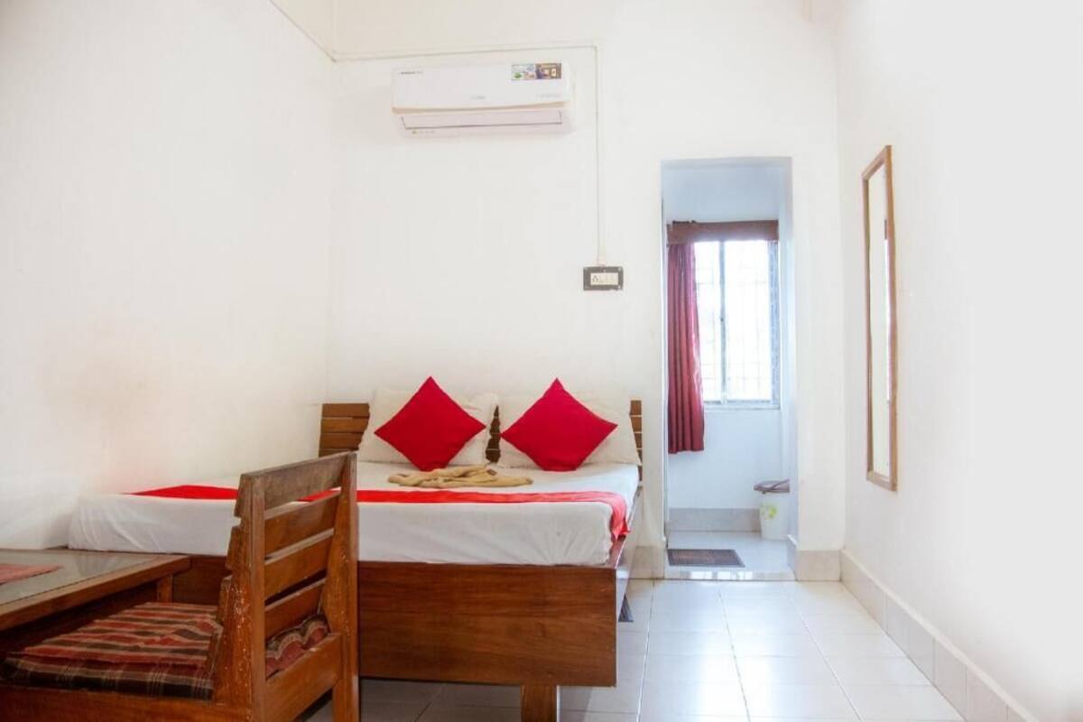 A room with a bed, a wooden chair, a small table, an air conditioning unit, and a door leading to another room.