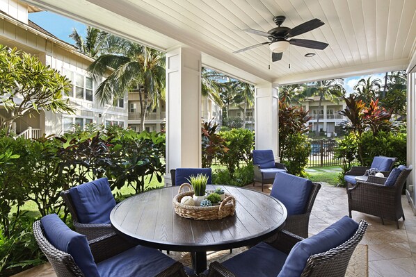 A covered outdoor seating area with wicker furniture and a central table, surrounded by lush greenery and a view of a residential building.