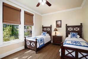 A bedroom with two wooden beds, a ceiling fan, a nightstand with a lamp, and a window with a view of greenery.