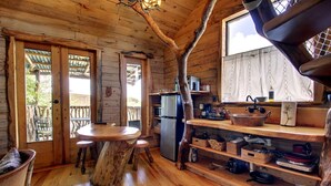 Una cabaña rústica de madera con comedor que incluye una mesa y bancos hechos con troncos de árbol, una cocina con electrodomésticos básicos y un ventanal con vista a una terraza.