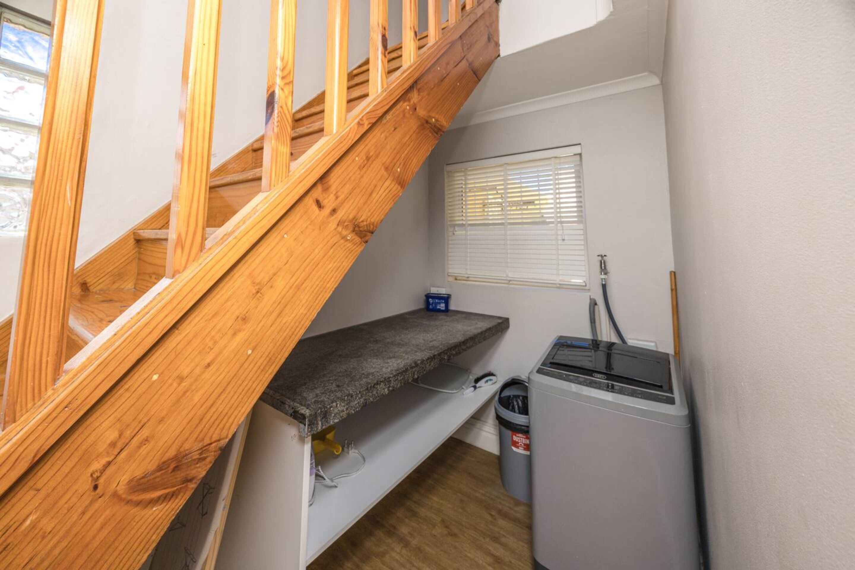 Une pièce de taille modeste avec un escalier en bois, une petite fenêtre avec des stores, une étagère intégrée et un lave-linge.