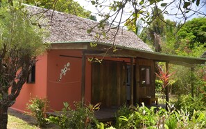 A small, single-story building with a thatched roof, surrounded by lush greenery and a variety of plants.