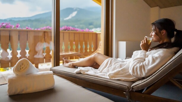 A person relaxing on a sun lounger with a view of mountains and flowers through a window. - Camina Suite & Spa (Cortina d'Ampezzo)
