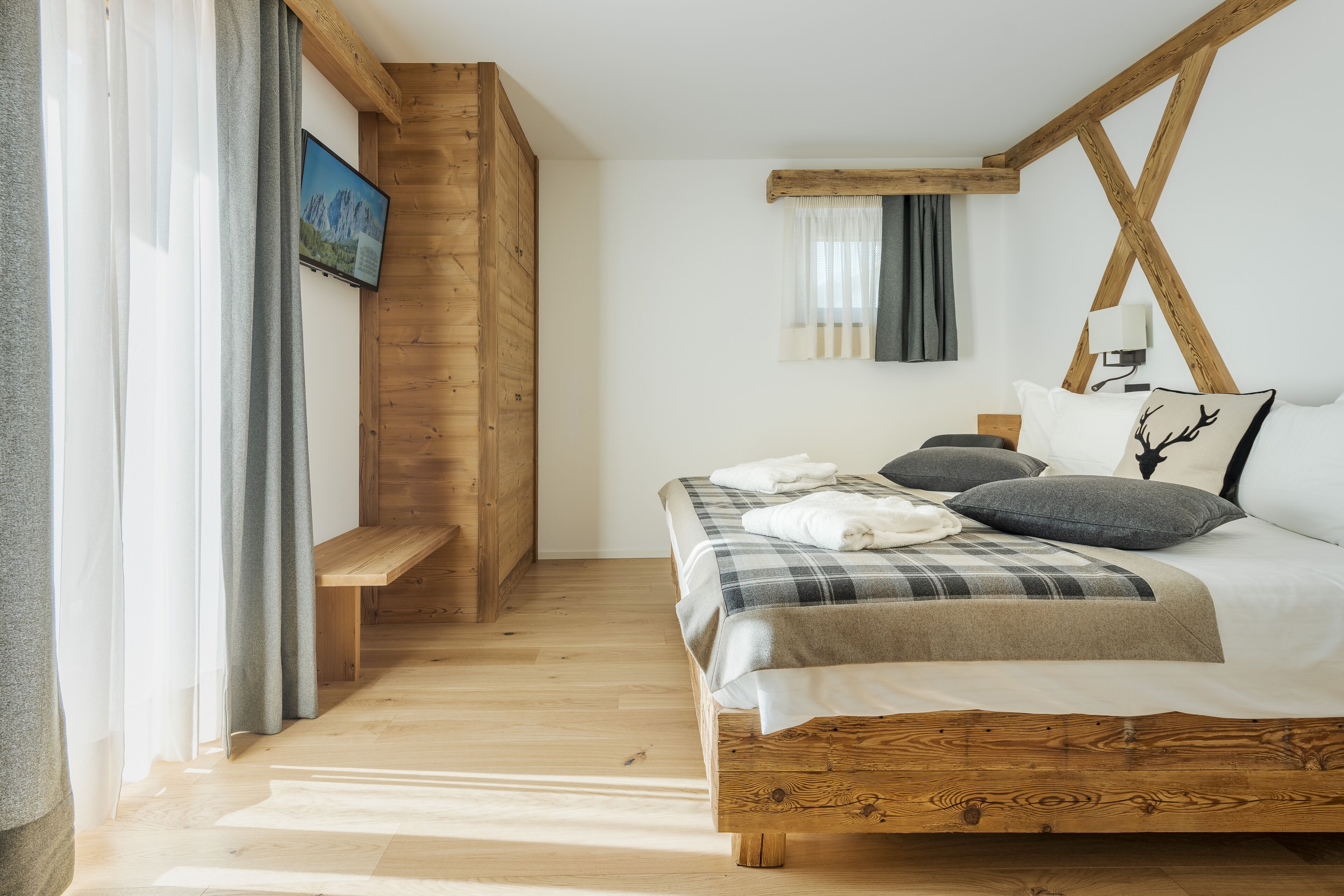 Ein Schlafzimmer mit einem hölzernen Bettgestell, einer Bank, einem an der Wand befestigten Fernseher und einem Fenster mit Vorhängen.