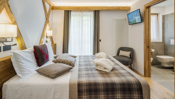 A hotel room with a bed, a chair, a television, and a bathroom visible through an open door. - Camina Suite & Spa (Cortina d'Ampezzo)