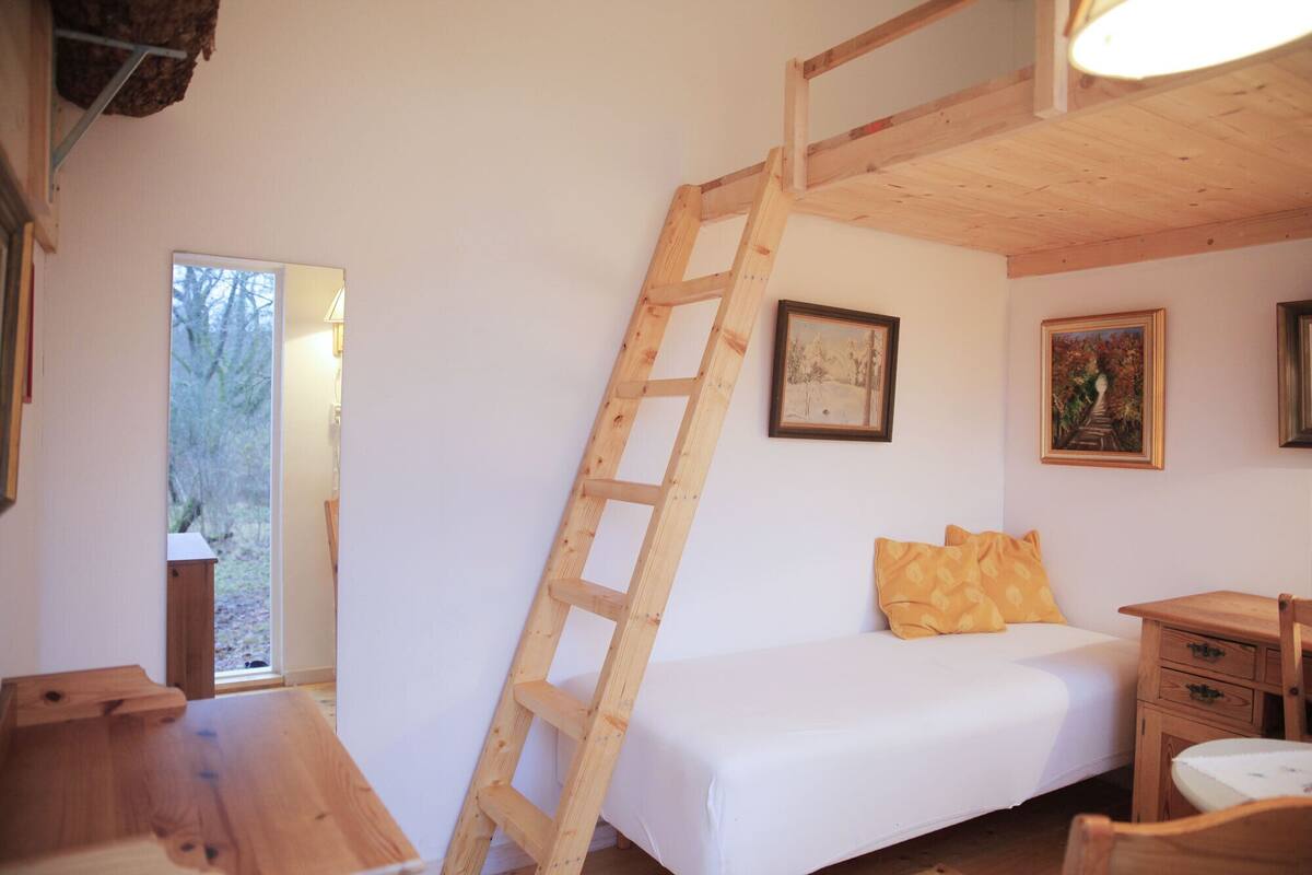 A bedroom with a wooden ladder, a bed, a desk, and two paintings on the wall.