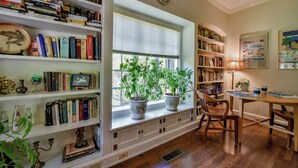 Una habitación con suelo de madera, una ventana con persianas, una estantería llena de libros, un reloj en la pared y una mesa con sillas.