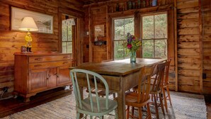 A wooden cabin interior with a dining table, chairs, a lamp, a vase with flowers, and a window with a view of trees. - Charming lakefront cabin close to Acadia National Park (Franklin)