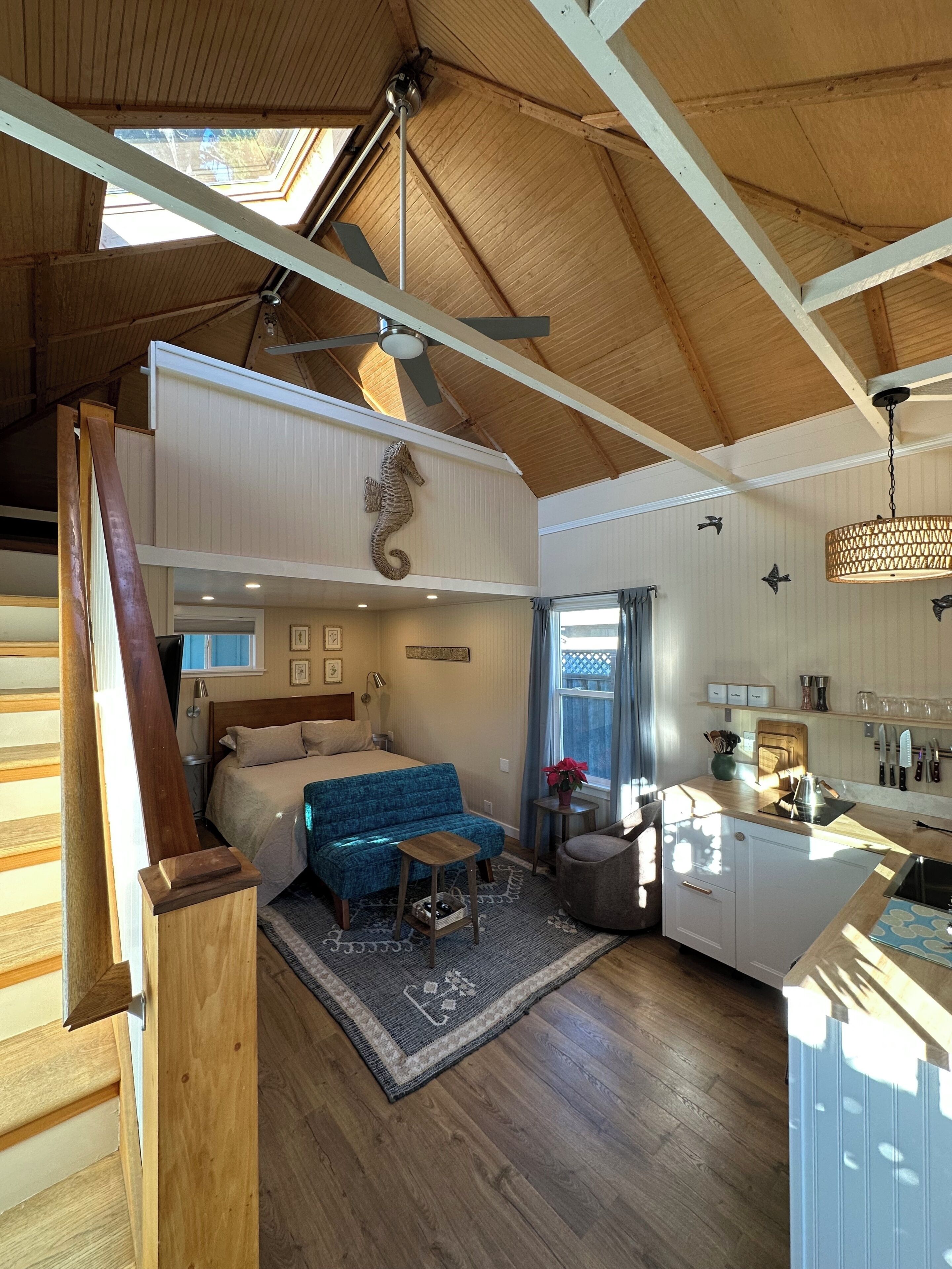 A spacious loft-style room with a high ceiling, a wooden staircase, a bed with a blue upholstered headboard, a kitchen area with a wooden countertop, and a skylight.