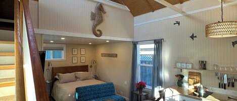 A spacious loft-style room with a high ceiling, a wooden staircase, a bed with a blue upholstered headboard, a kitchen area with a wooden countertop, and a skylight.