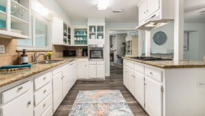 A modern kitchen with white cabinetry, granite countertops, and a central island. There are built-in appliances, including a microwave and oven, and a window with blinds. - POOL House on the Kemah Boardwalk w BIKES (Kemah)