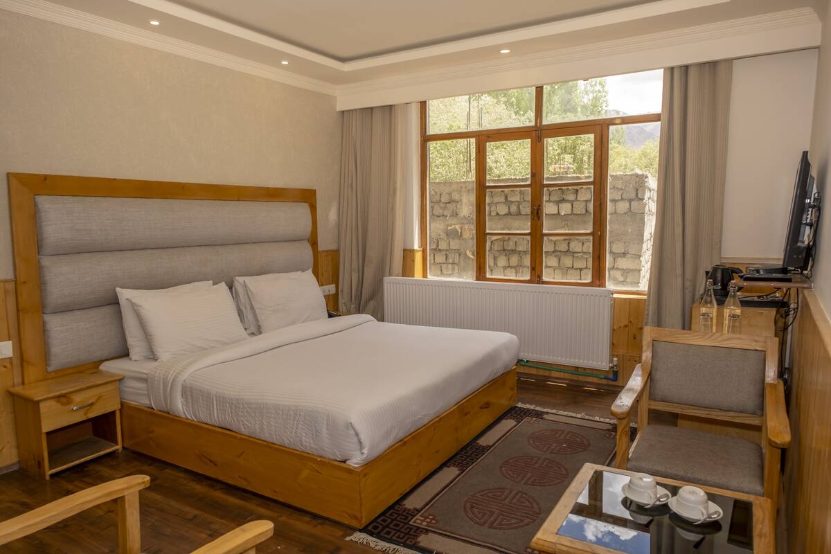 A bedroom with a large bed, wooden headboard, a nightstand, a wooden chair, a small table with cups, and a window with a stone wall view.