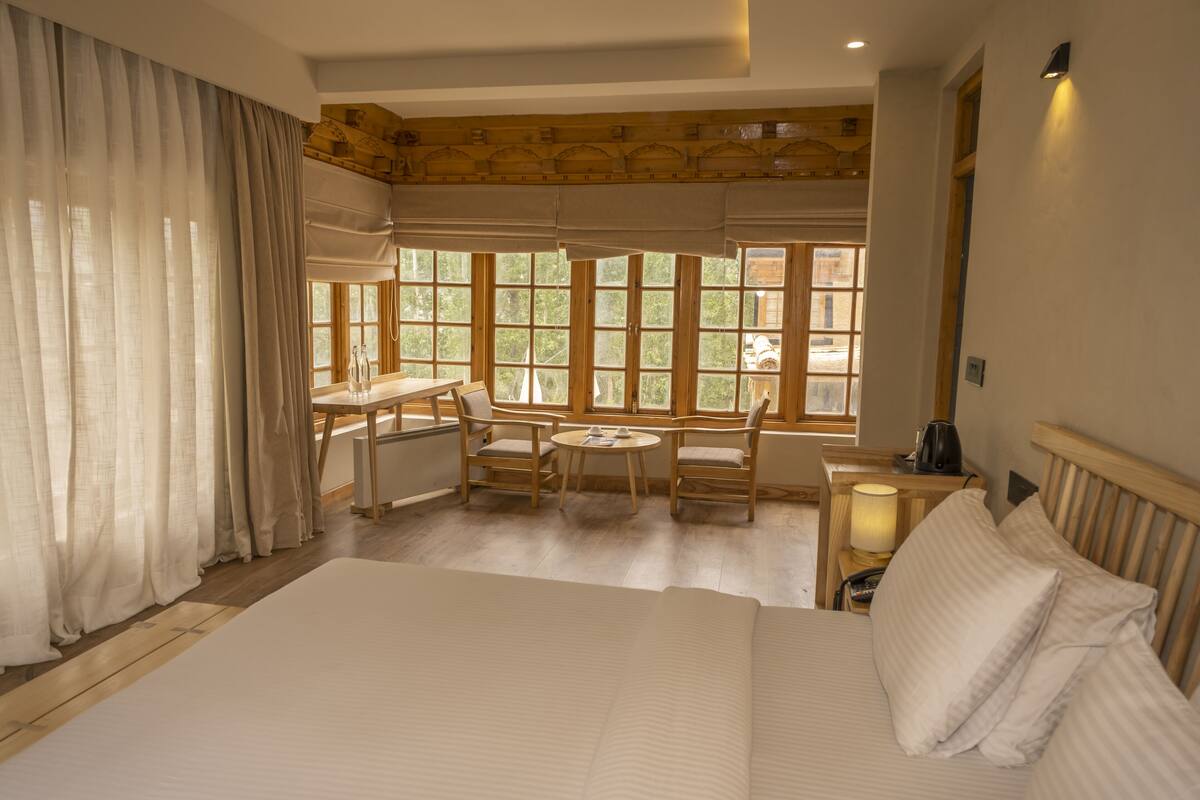 A bedroom with a large bed, a desk with chairs, and a window with a view of trees.