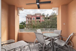 Un balcon avec une table et des chaises, un ventilateur de plafond et une vue sur un grand bâtiment à l’architecture rouge et blanche.