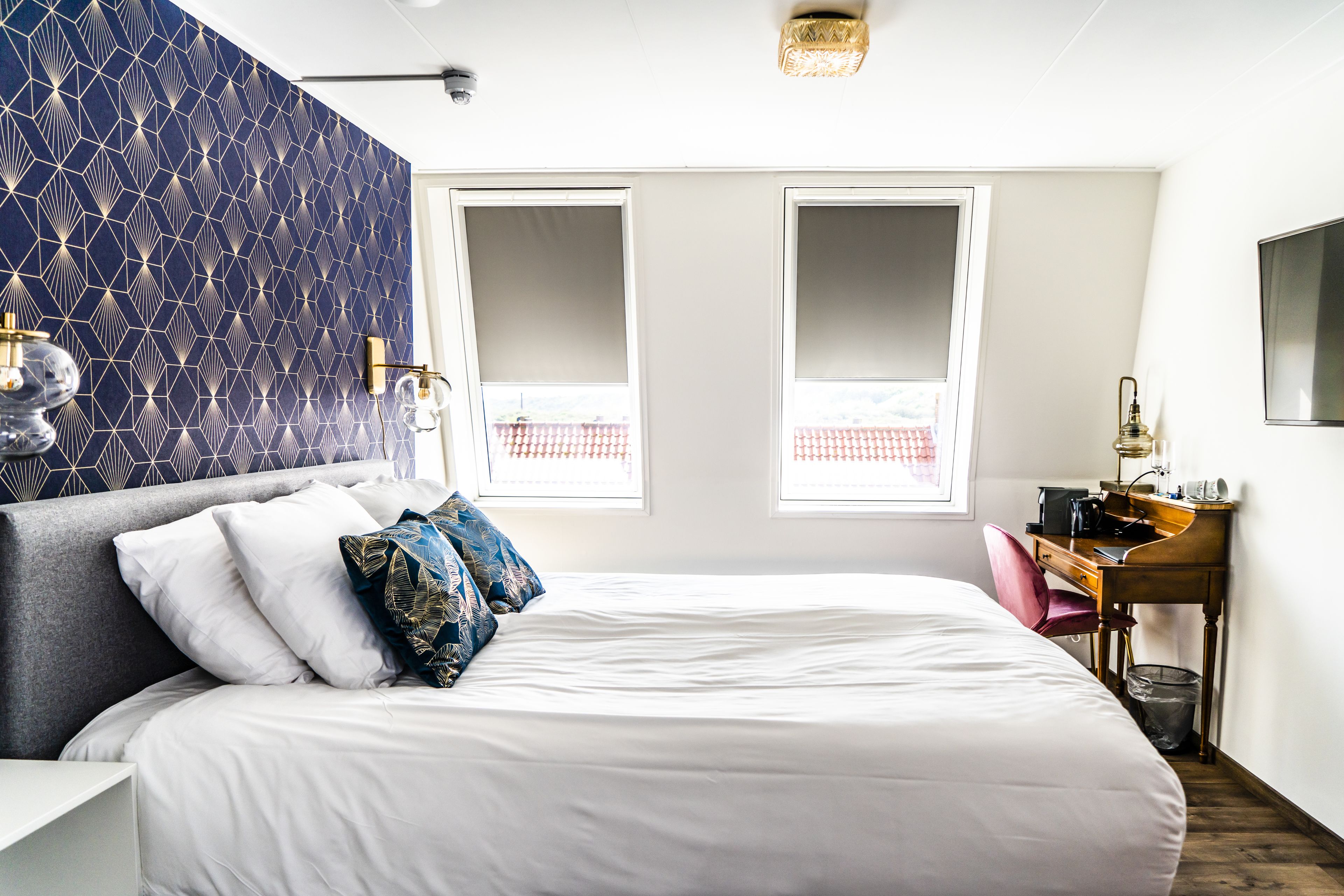 A neatly arranged bedroom with a large bed, a desk, and a chair, featuring a geometric patterned wall and a window with a view.