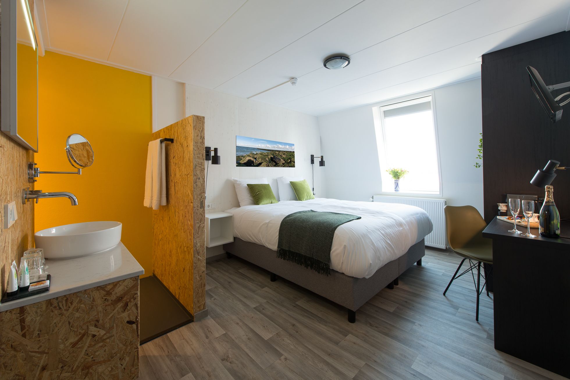 Quarto moderno com uma cama grande, mesa de cabeceira, janela com vista, banheiro com pia e espelho, e escrivaninha com cadeira.