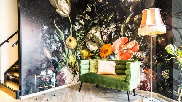 Una habitación con un sofá verde, una lámpara de pie y un mural floral en la pared.