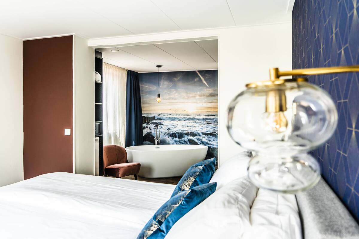 A hotel room with a large bed, a bathtub, a chair, and a view of the ocean through a mural.