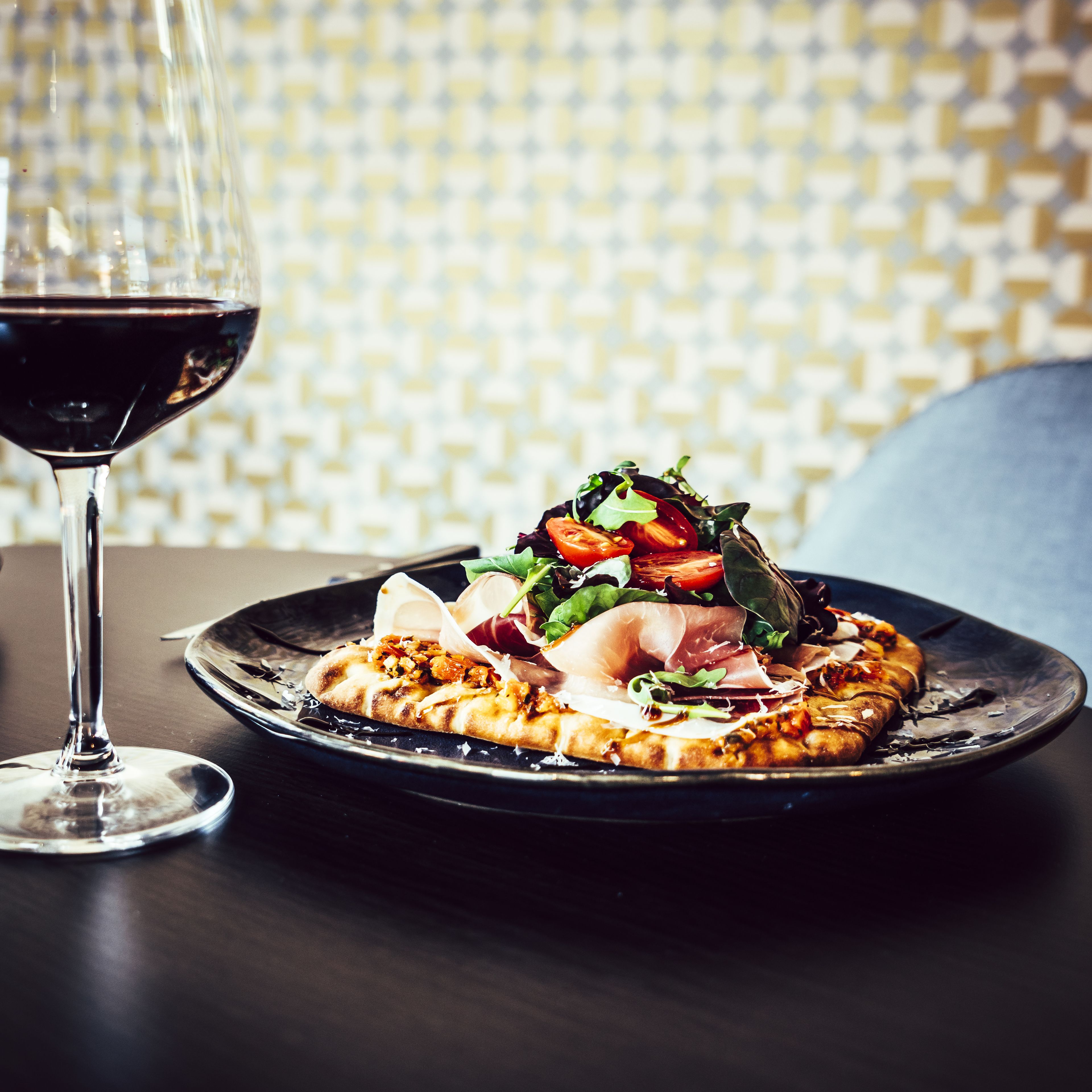 Una pizza con diversi condimenti su un piatto nero, un bicchiere di vino rosso e un muro decorato sullo sfondo.