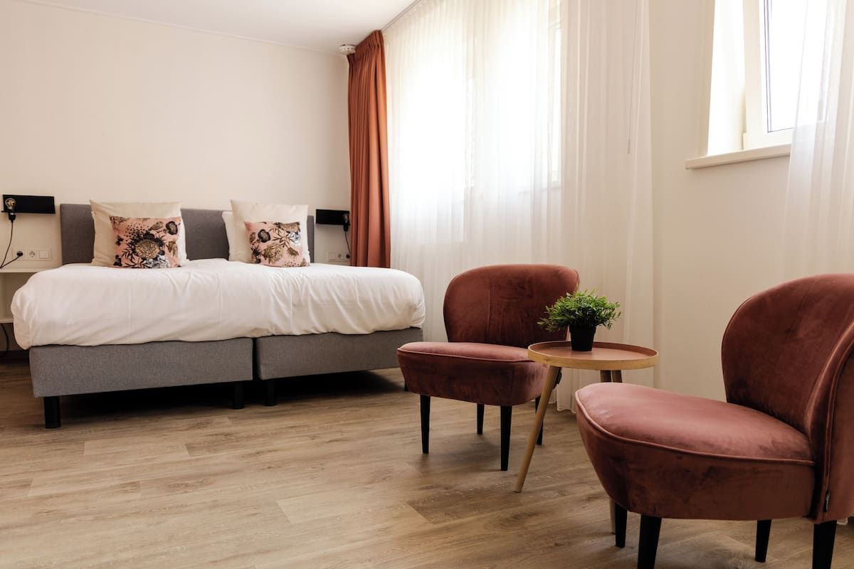 A modern hotel room with a grey sofa bed, two red armchairs, a small round table with a plant, and a window with sheer curtains.