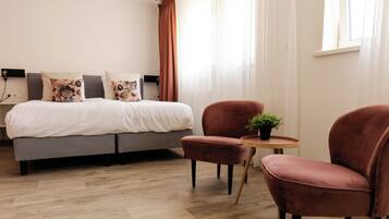Quarto de hotel moderno com sofá-cama cinza, duas poltronas vermelhas, uma mesa redonda pequena com uma planta e uma janela com cortinas leves.