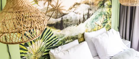 A bed with white and grey pillows, a woven hanging light fixture, and a tropical mural on the wall.