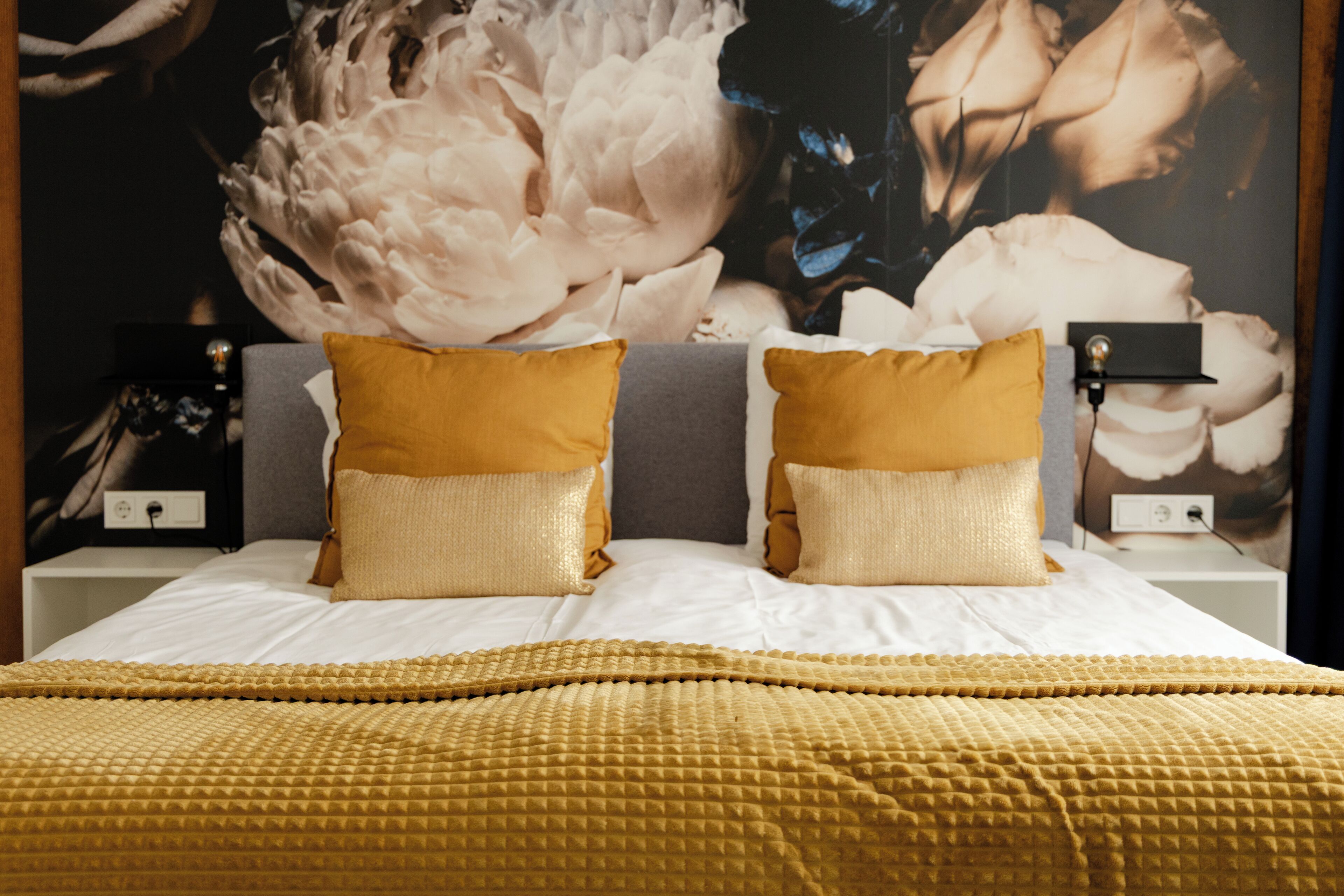 A bed with a gold textured blanket and pillows, a large floral artwork on the wall, and bedside tables with lamps.