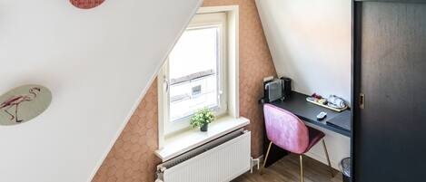 A small, triangular attic bedroom with a window, radiator, desk, chair, and a door.