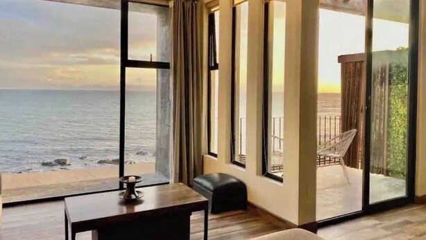 A room with a large window overlooking the sea, a sliding glass door to an outdoor area, and a small table with a candle. - Ensenada’s Luxury Ocean Front Property. 1or 2 condominiums available. (Ensenda)