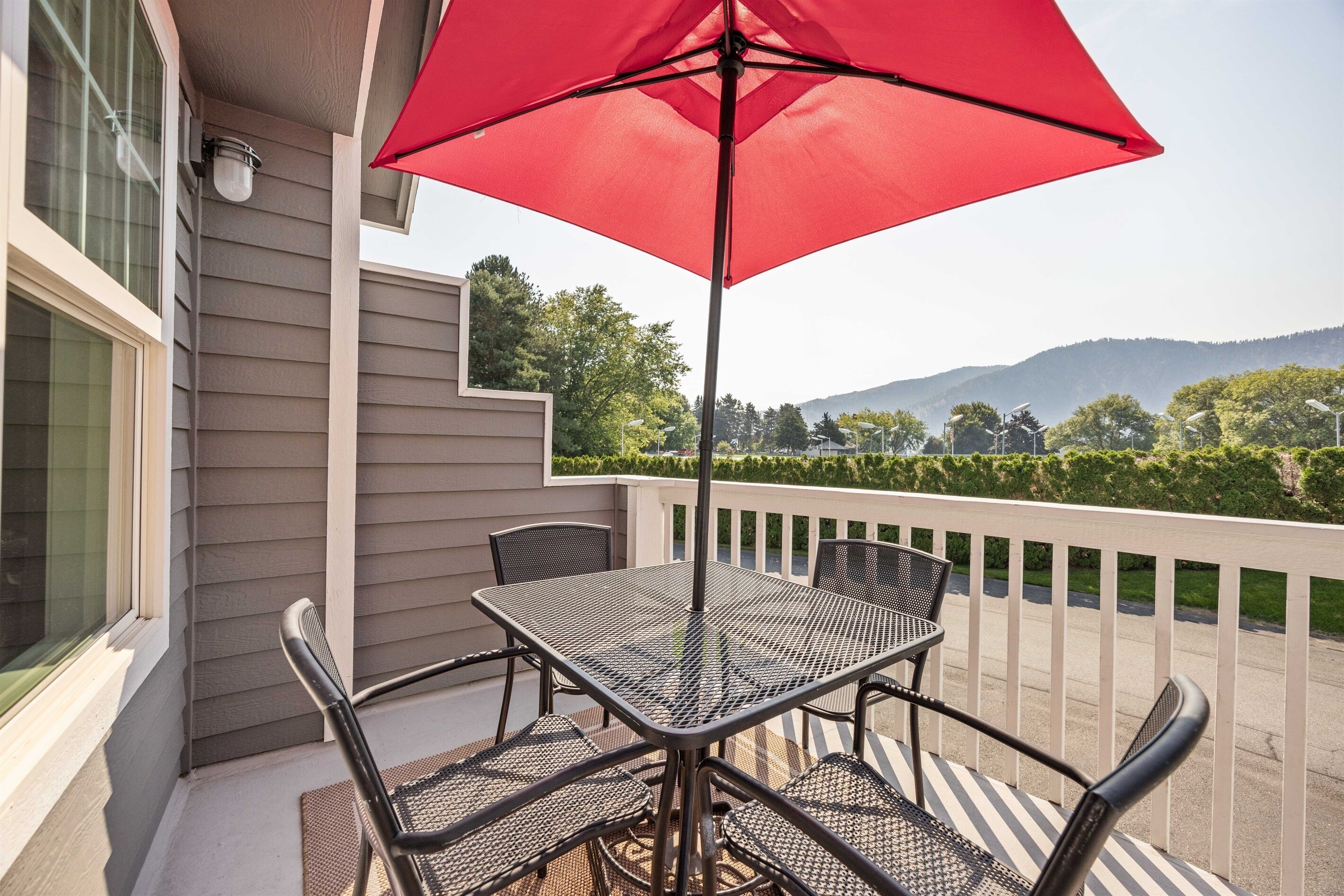 Un balcon avec une table et des chaises, un parasol rouge, et une vue sur les arbres et les collines.