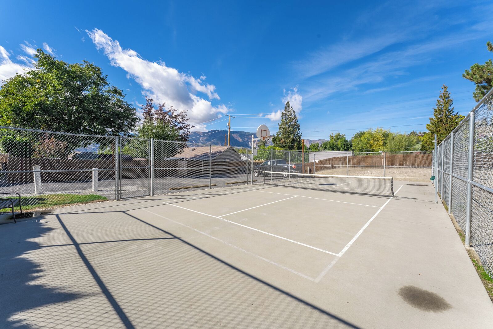 Ein Tennisplatz im Freien mit Netz, umgeben von Maschendrahtzäunen, und ein Basketballkorb im Hintergrund.