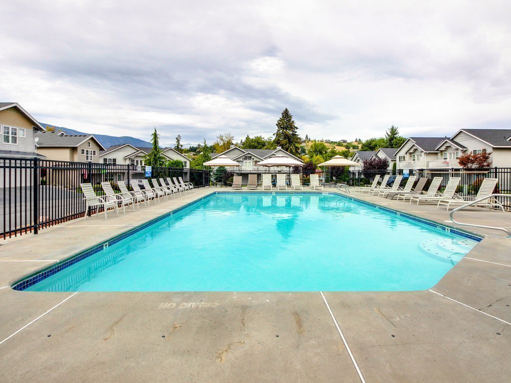 Une piscine entourée de chaises longues et d’une clôture, avec des maisons en arrière-plan.