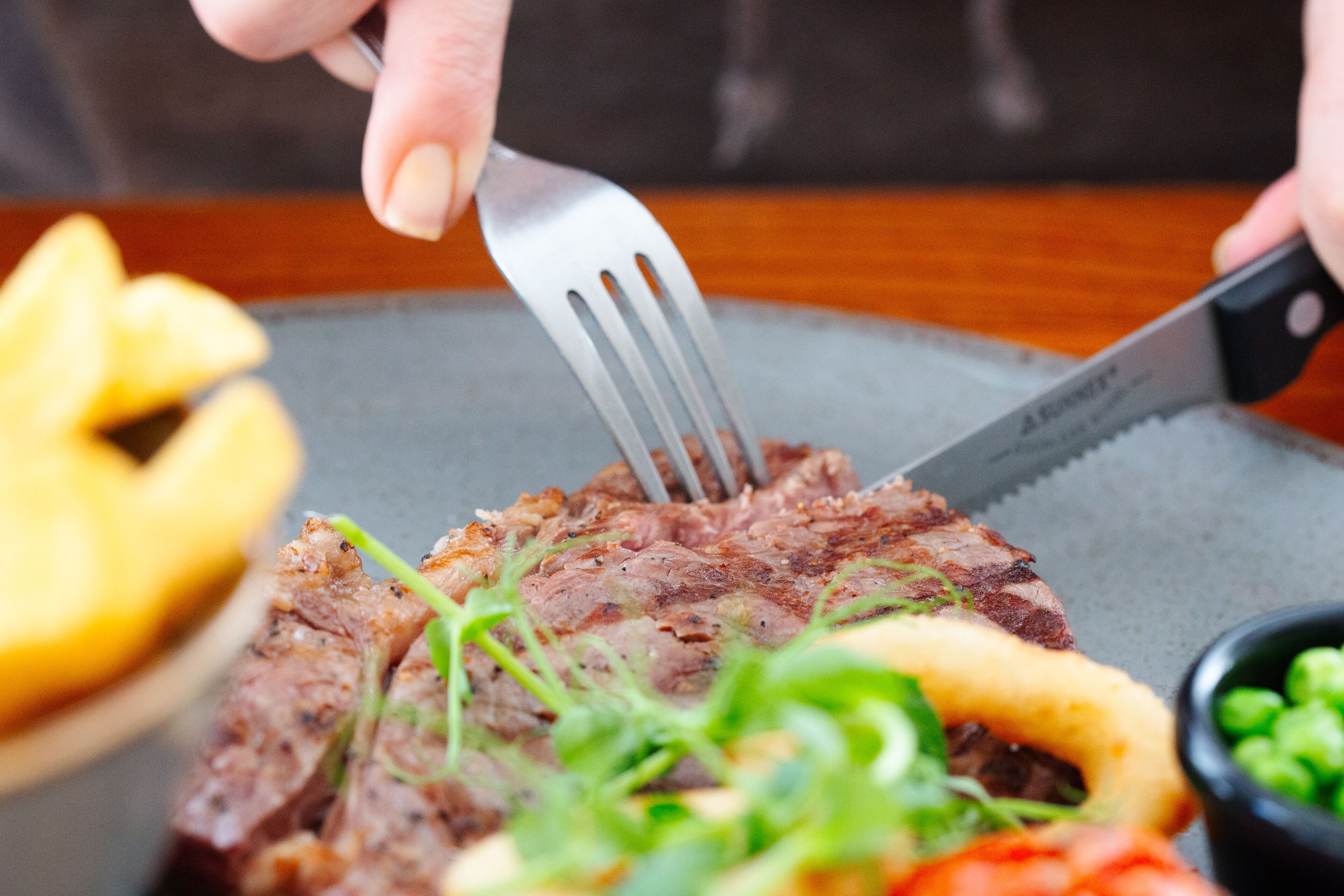 A close-up of a steak with a fork and knife on a plate, accompanied by a side of greens and a small bowl of peas.