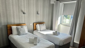 Une chambre d’hôtel avec deux lits simples, chacun doté d’une tête de lit en bois et d’un revêtement gris, ainsi que d’une literie blanche. On y trouve une fenêtre avec des rideaux, un radiateur et une chaise dans un coin.