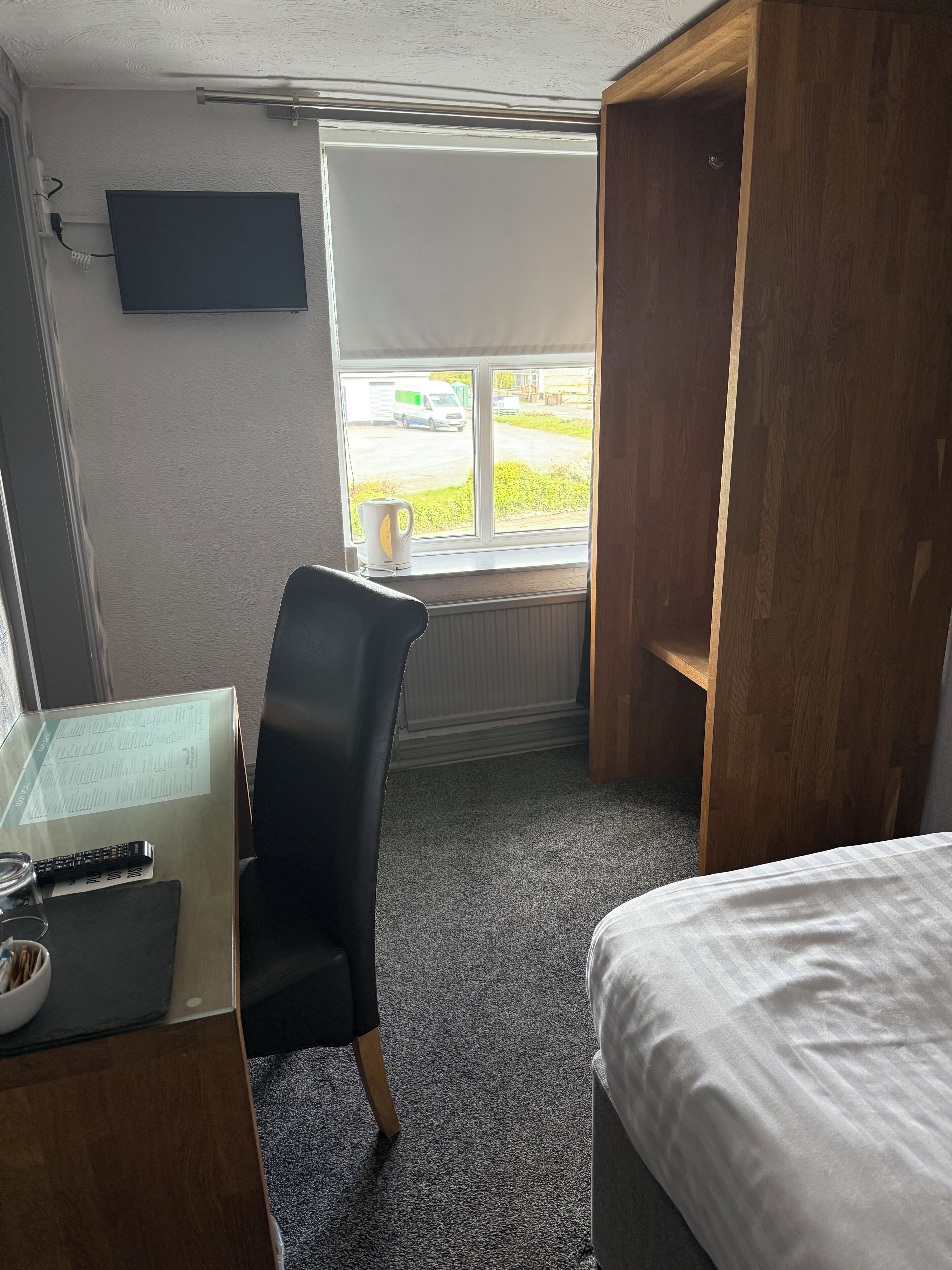 Una habitación de hotel con cama, escritorio con computadora, silla, ventana con vista a un estacionamiento y un radiador.