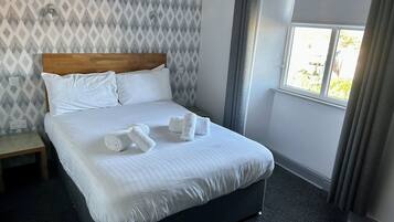 A neatly made bed with white linens and two rolled towels, a wooden headboard, a nightstand with a lamp, and a window with curtains.