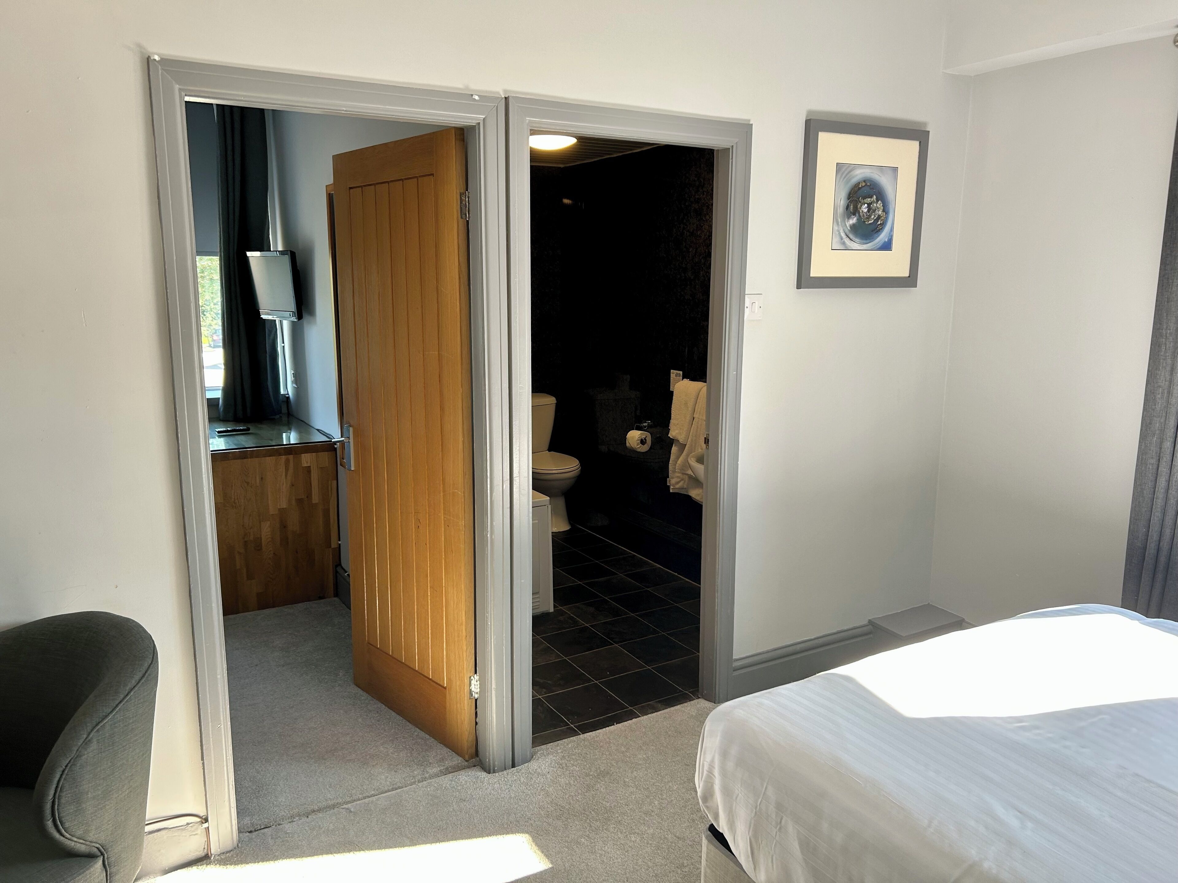 Una habitació d'hotel amb un llit, una porta de fusta que dona a un bany, un quadre amb marc a la paret i un escriptori petit amb una cadira.