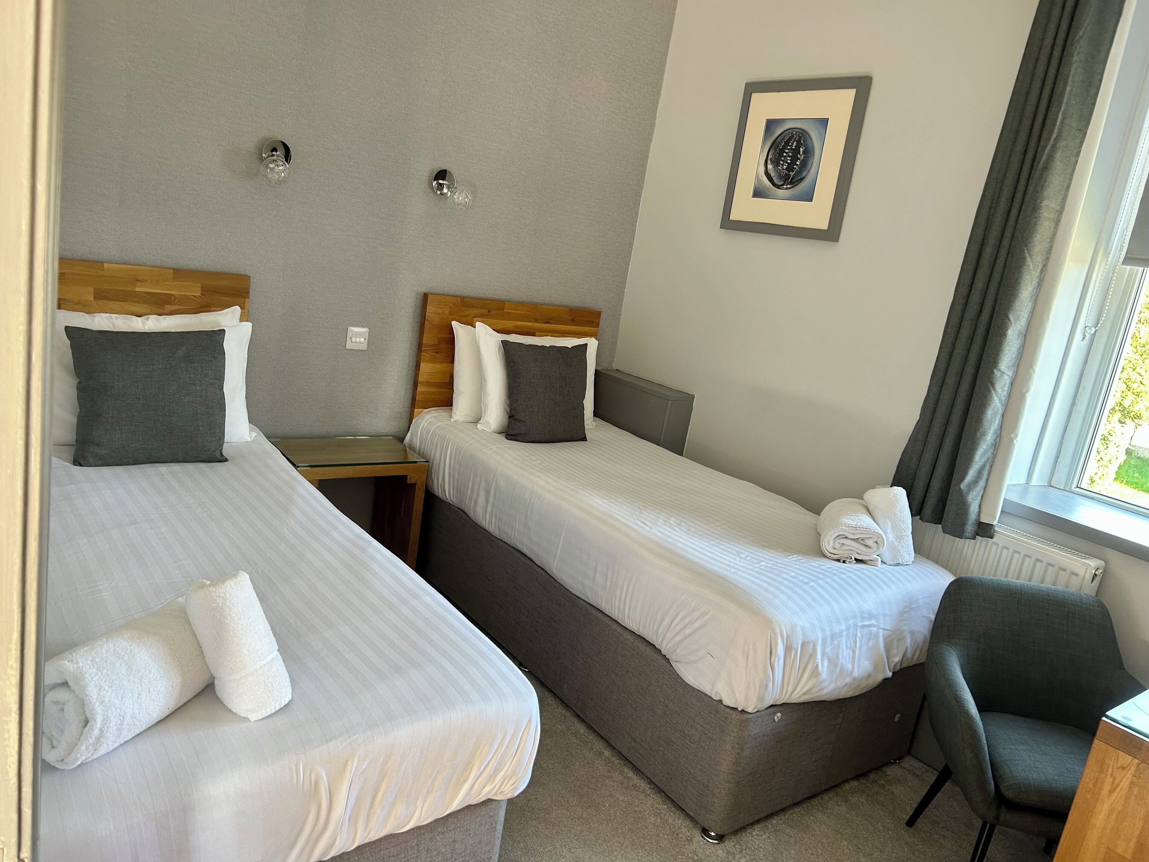 A hotel room with two single beds, a small wooden nightstand, a grey armchair, and a framed picture on the wall.