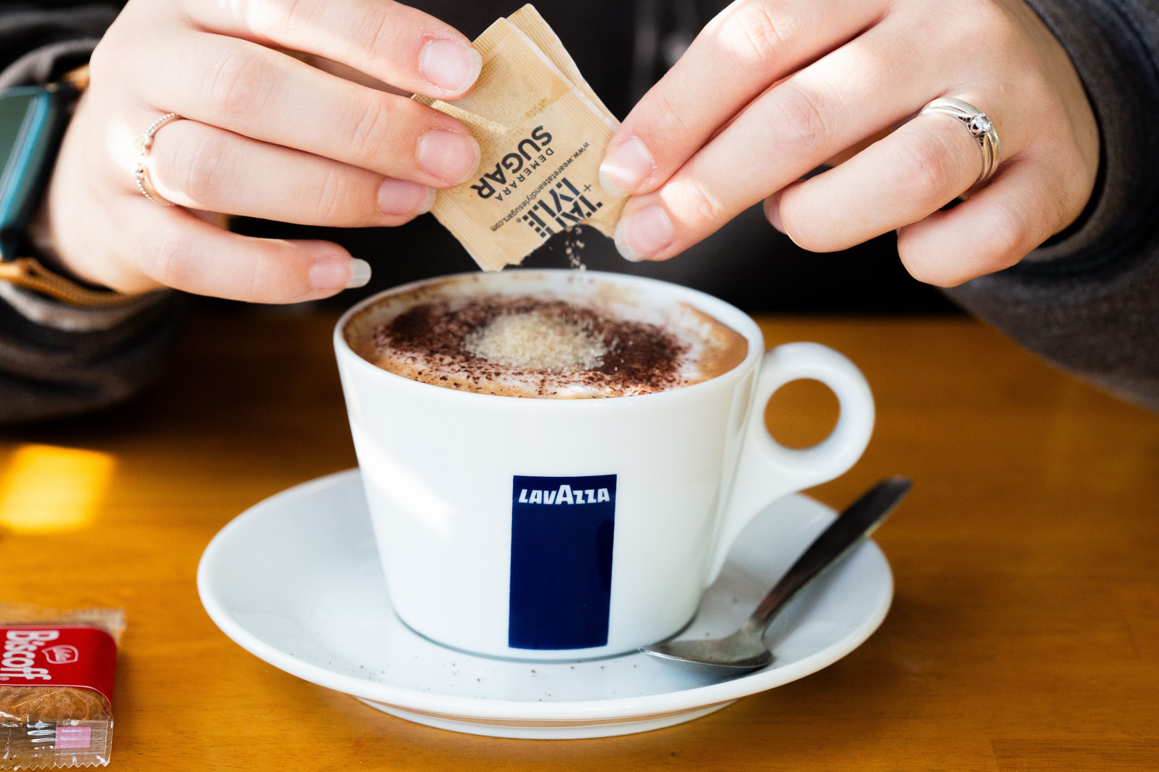 A cup of coffee with a spoon on a saucer, a packet of sugar, and a biscuit packet on a table.