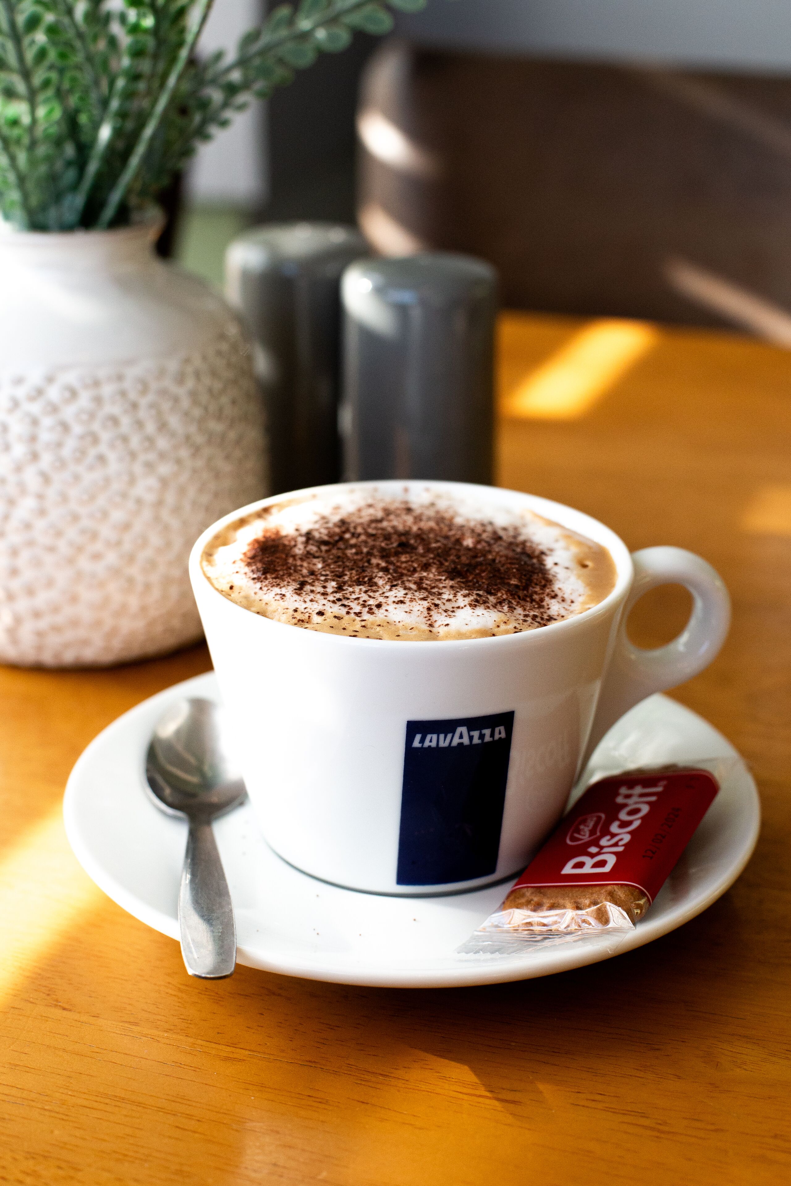 A cup of coffee with a chocolate topping, a biscuit packet, and a spoon on a saucer.