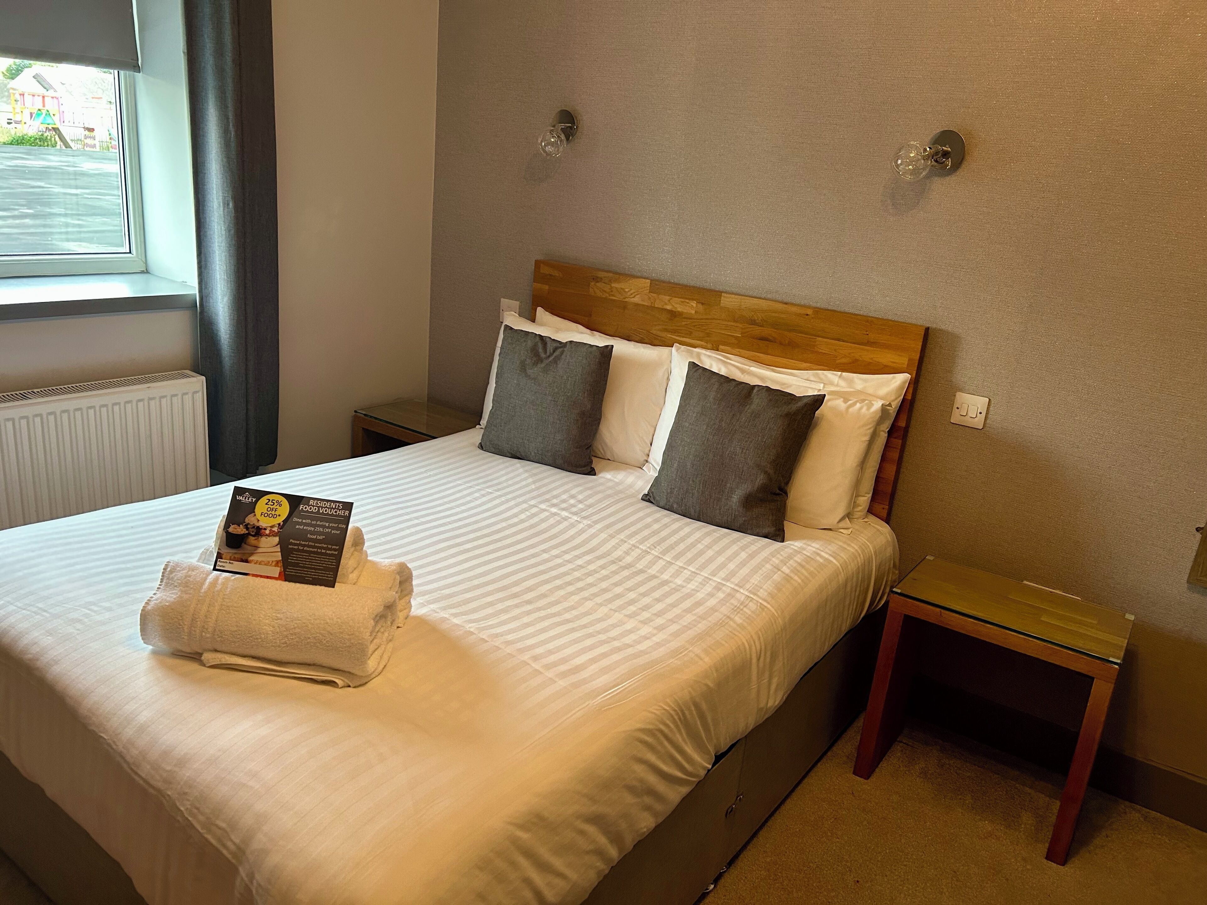 A neatly made bed with a folded towel and a food voucher, a bedside table, and a nightstand.