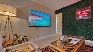 Ein Wohnzimmer mit großem Flachbildfernseher, einer grünen Wand mit einer Neonreklame, einem Holztisch und einer Lampe.