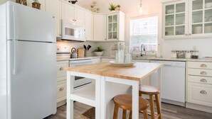 Une cuisine moderne avec des meubles blancs, un îlot central doté de tabourets, un réfrigérateur, un four à micro-ondes et une fenêtre laissant entrer la lumière naturelle.