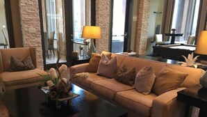 Una sala de estar con sofá color beige, una mesa de centro y una lámpara. Se ve el exterior a través de ventanas amplias.