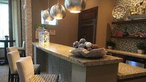 Una cocina con encimera de granito, dos taburetes de bar, iluminación colgante y objetos decorativos en los estantes.