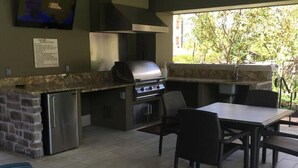 Cocina exterior con una parrilla, un refrigerador y un comedor con sillas y una mesa.