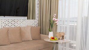A living room with a beige sofa, a small round table with a vase of flowers, a fruit bowl, and a lamp. - Europe Hotel & Casino (Sunny Beach)